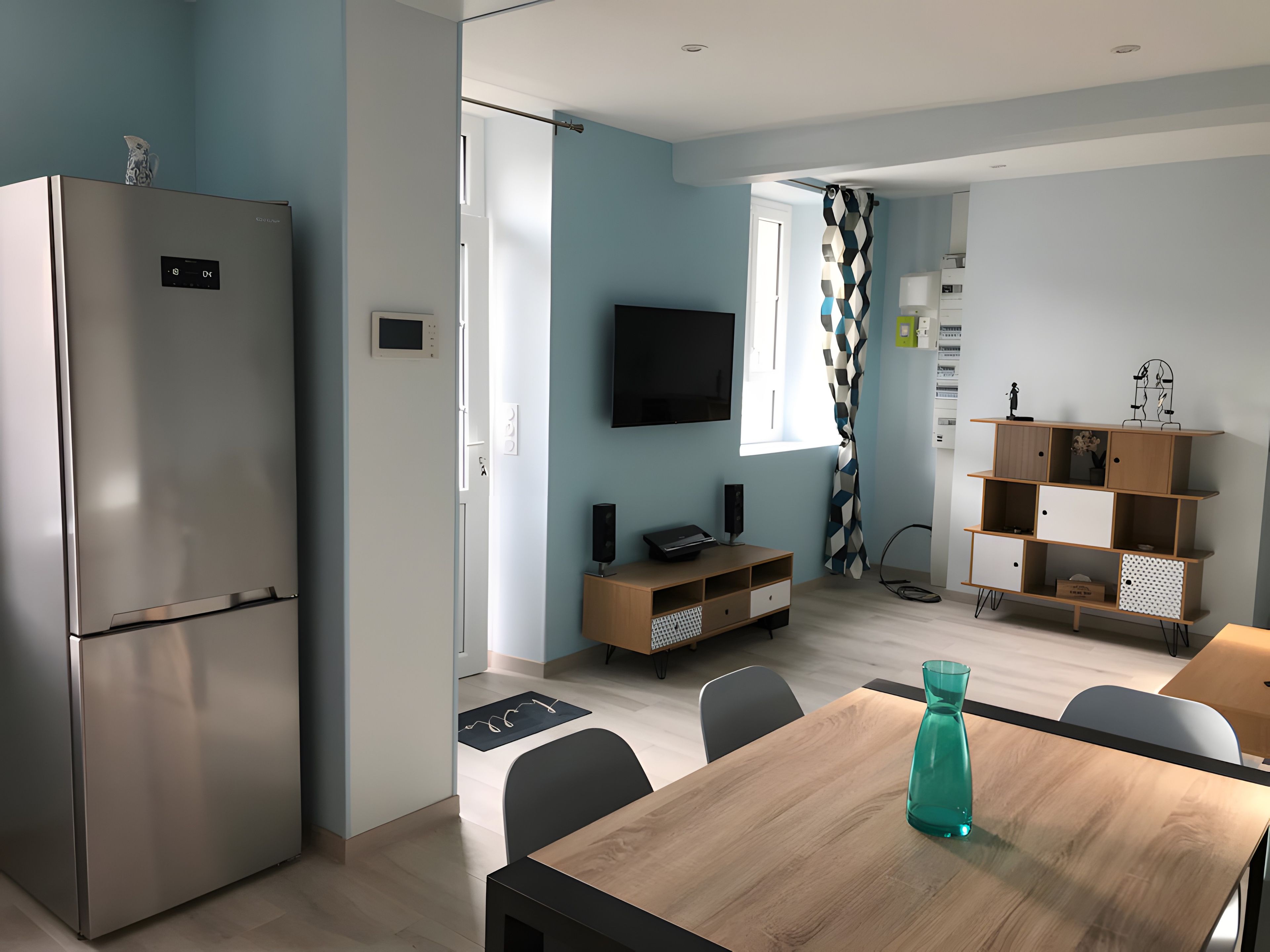 A modern kitchen with a built-in refrigerator, a dining table with chairs, a TV mounted on the wall, and a patterned curtain.