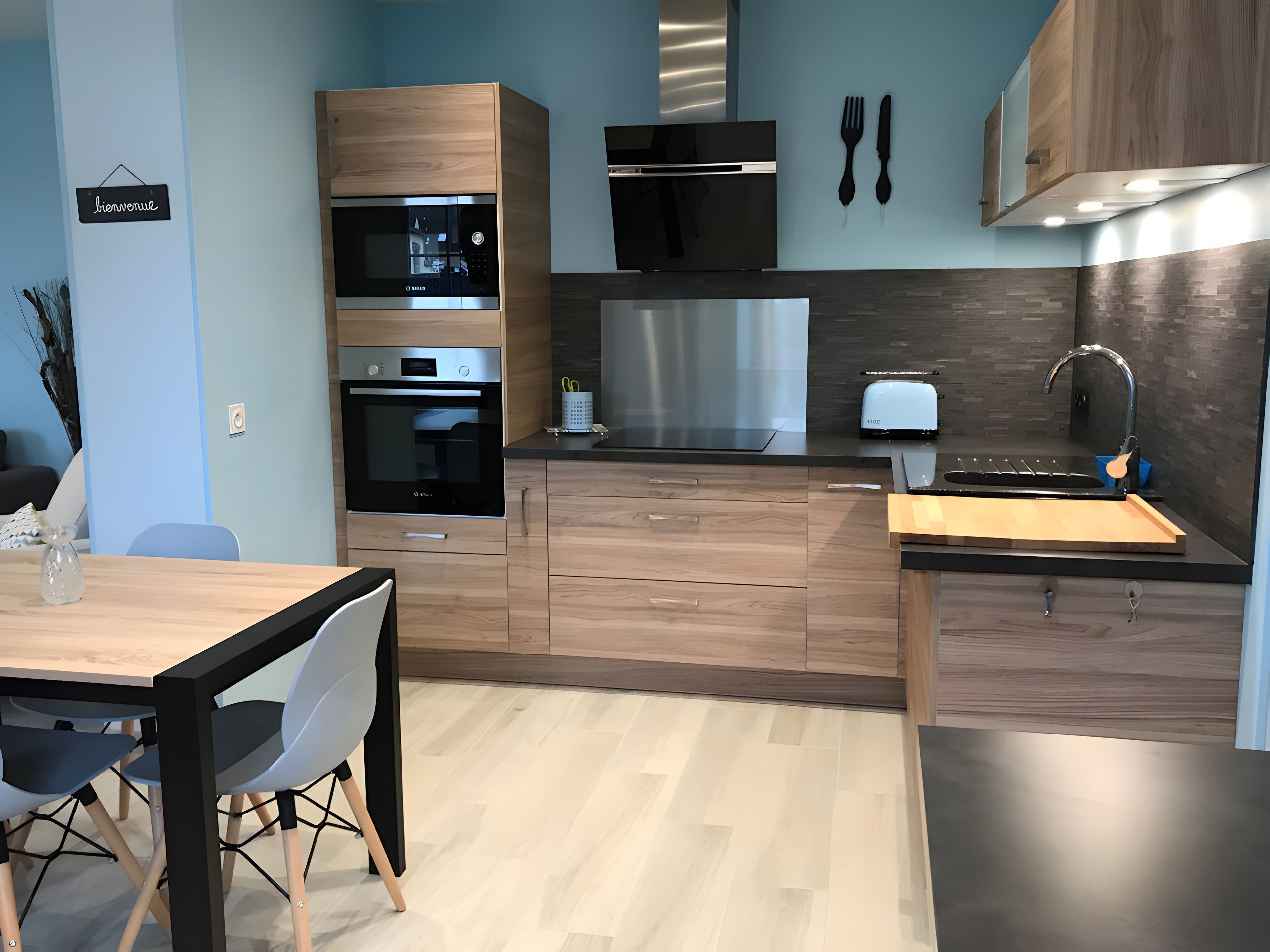 Una cucina moderna con elettrodomestici integrati, una zona pranzo con tavolo e sedie, e un cartello con la scritta 'Bienvenue' al muro.