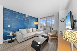 A modern living room with a large window, a sofa, a coffee table, and a map wall. - Amazing Apt w Bay View Balcony - Near Beach (Hallandale Beach)