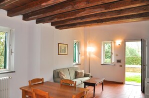 A room with a wooden ceiling, a sofa, a table, and chairs. There are windows and a door.