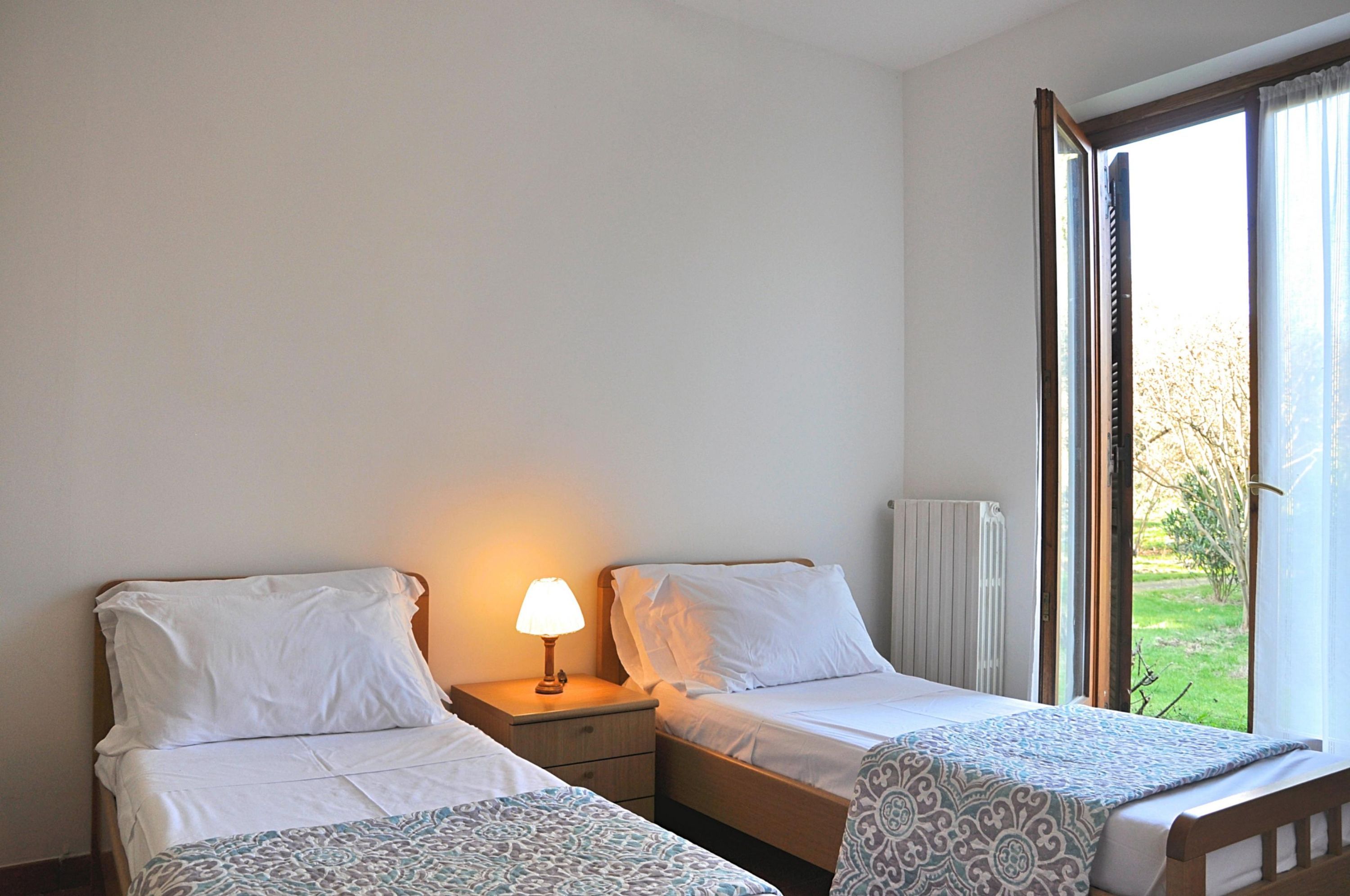 Et hotelværelse med to enkeltsenge, et natbord med lampe, en radiator og et vindue med gardiner.