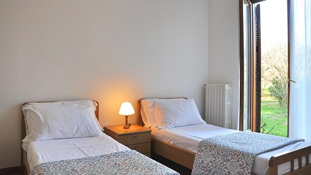 A hotel room with two single beds, a nightstand with a lamp, a radiator, and a window with curtains. - Montecasciano Mimosa (Capranica)
