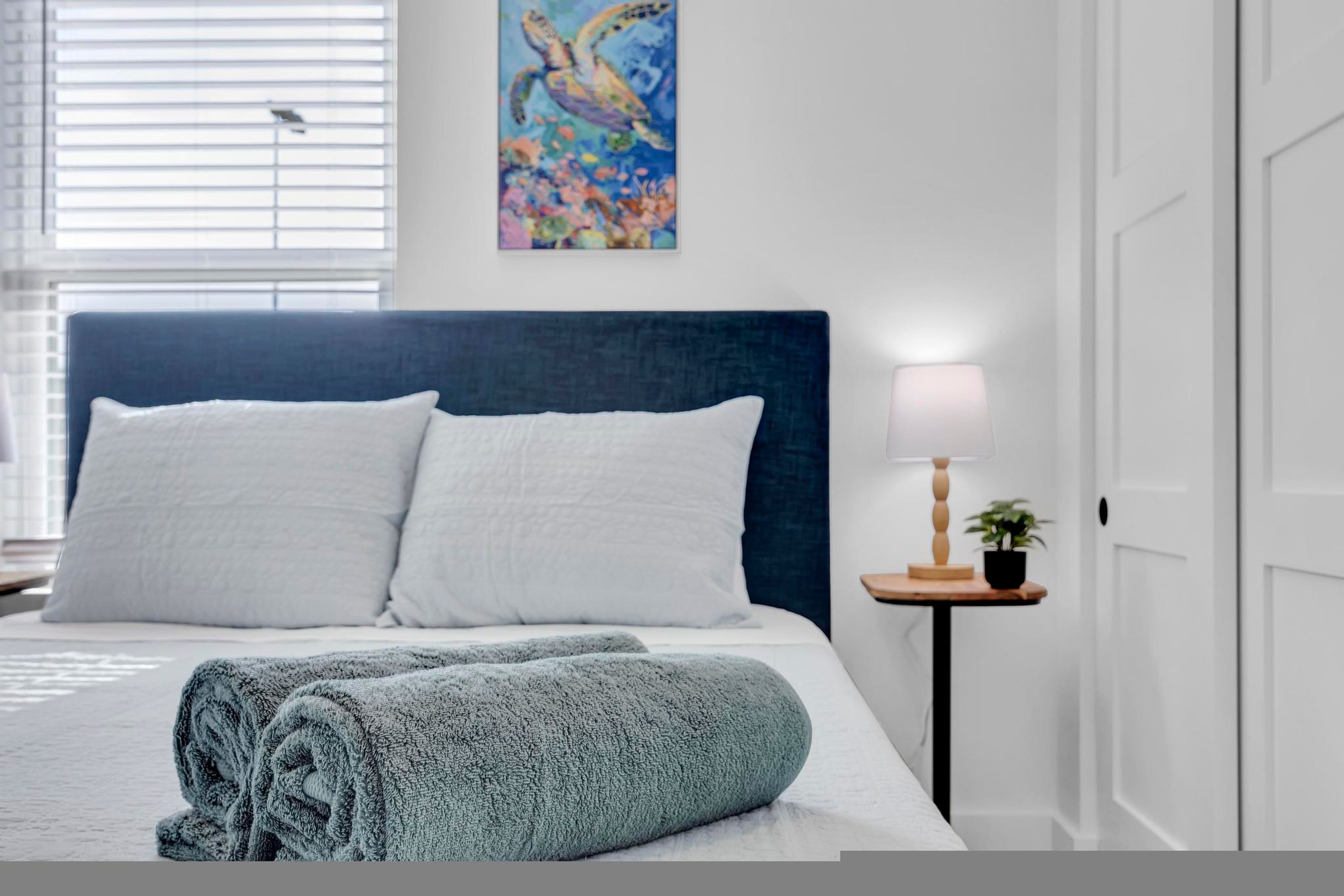Un lit bien fait, avec du linge de lit blanc et bleu, une tête de lit bleue et un tableau représentant une tortue de mer accroché au mur.