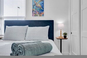 Una cama bien hecha con ropa de cama blanca y azul, un cabecero azul y un cuadro de una tortuga marina en la pared.