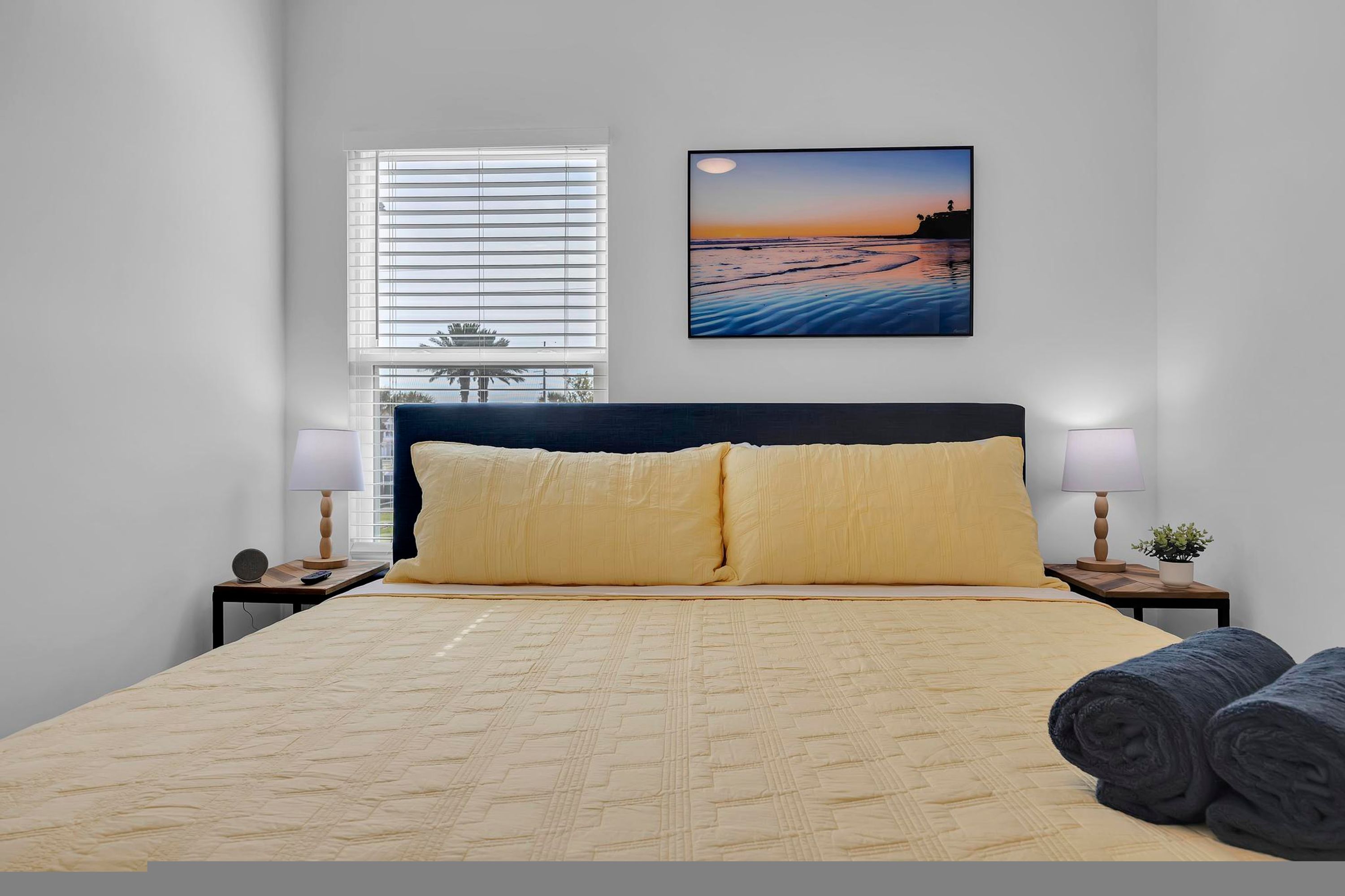 Une chambre à coucher comprenant un lit, deux tables de chevet, une commode, une fenêtre avec des stores, un tableau accroché au mur et une vue sur l’extérieur.