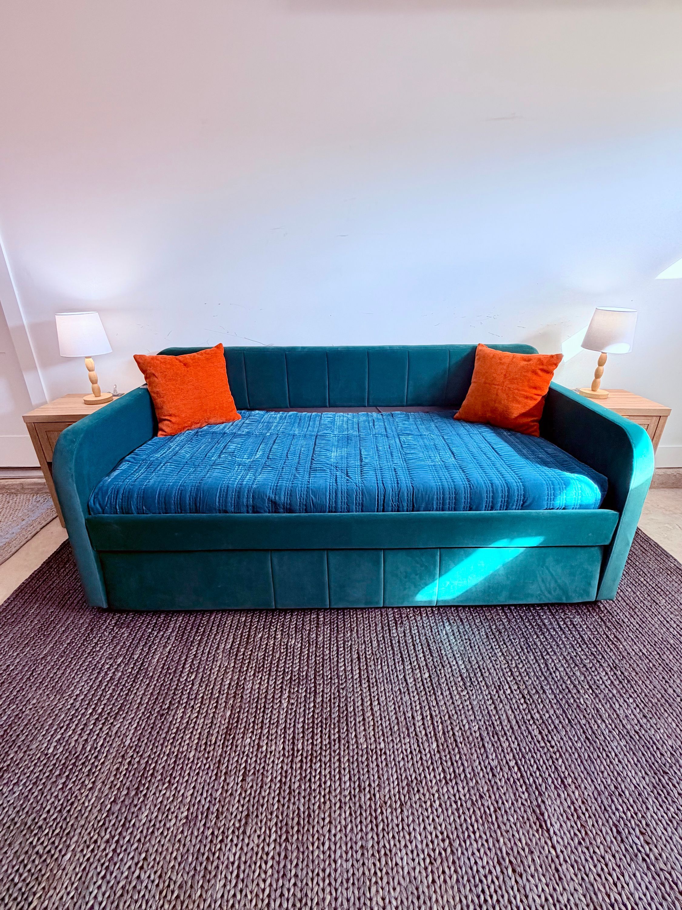 Un sofá color turquesa con cojines azules y naranjas en una habitación con alfombra tejida.