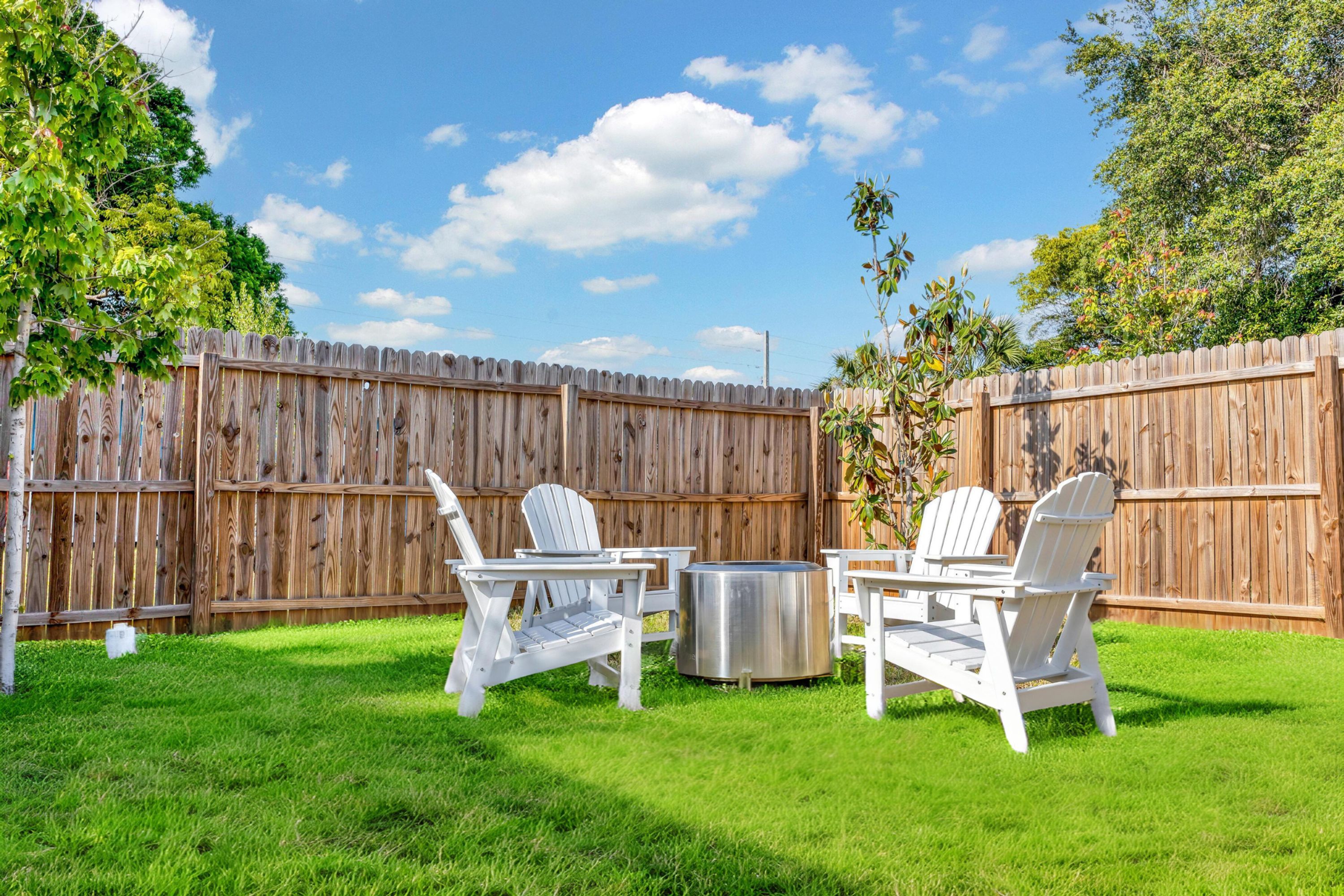 Un patio trasero con cerca de madera, dos sillas Adirondack blancas y una parrilla metálica.