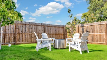 Un patio trasero con cerca de madera, dos sillas Adirondack blancas y una parrilla metálica.