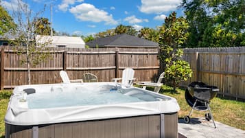 A backyard with a hot tub, two lawn chairs, a barbecue grill, and a wooden fence.