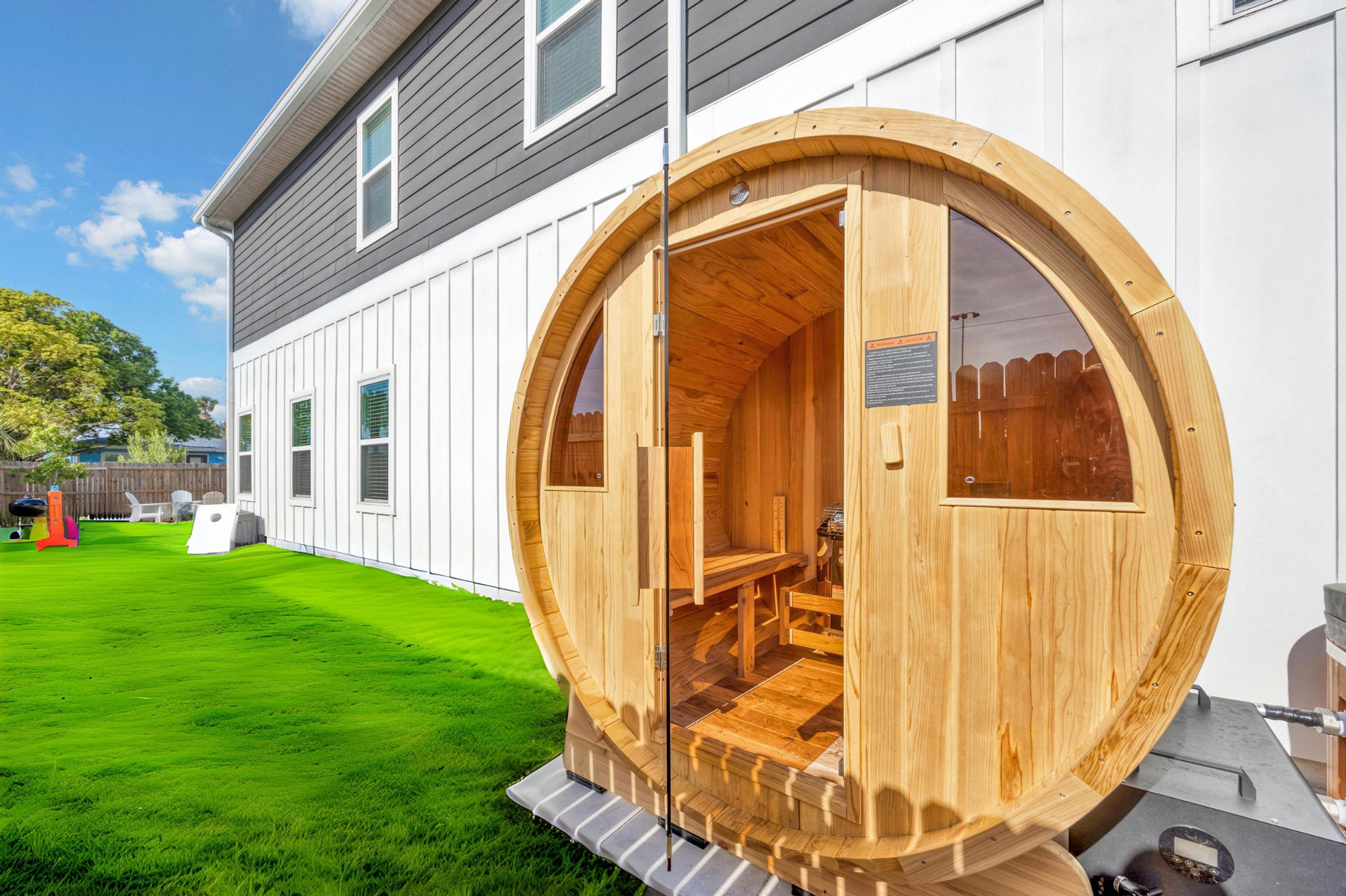 Una estructura de sauna de madera con un banco en el interior, adosada a una casa con revestimiento blanco y césped verde.