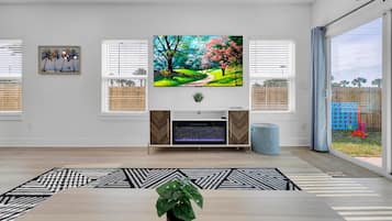 A living room with a flat-screen TV mounted above a fireplace, a dining table with a plant, and a view of the outside.