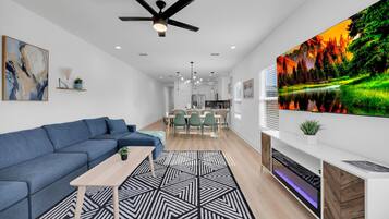 A modern living room with a blue sofa, a geometric rug, a ceiling fan, and a large TV displaying a nature scene.