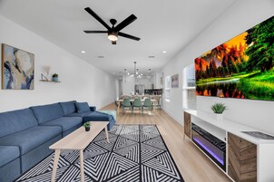 Uma sala de estar moderna com um sofá azul, um tapete geométrico, um ventilador de teto e uma televisão grande exibindo uma paisagem natural.