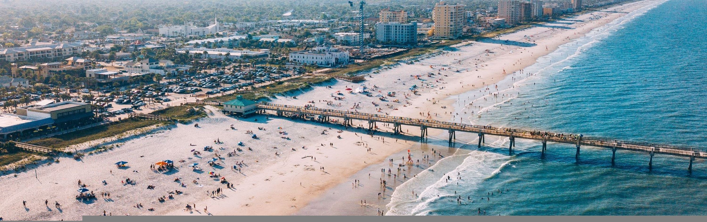 An aerial view of a beach with a pier, buildings, and a coastal cityscape.