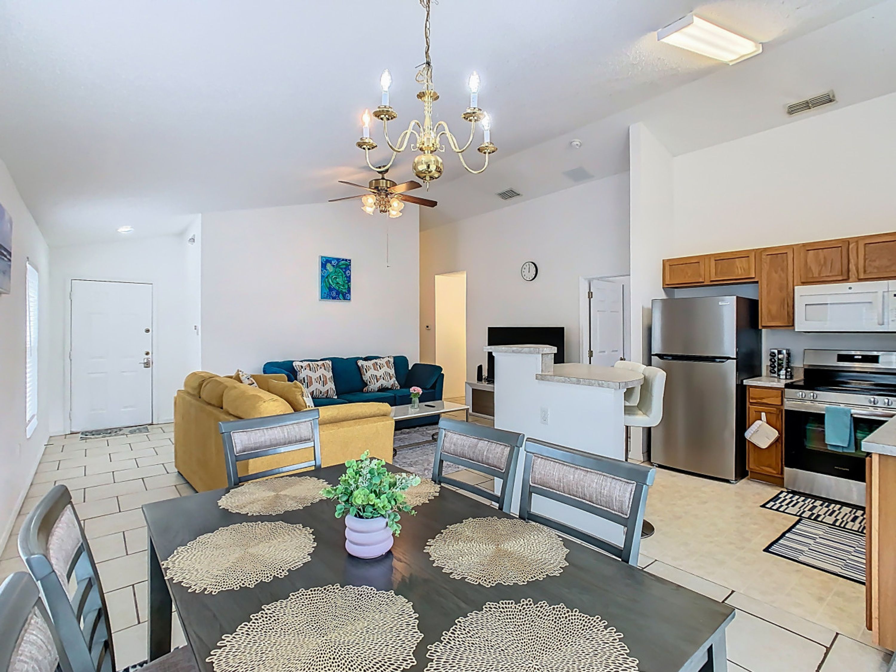 A modern kitchen with stainless steel appliances, a dining area with a table set for four, and a living room with a sofa and a TV.