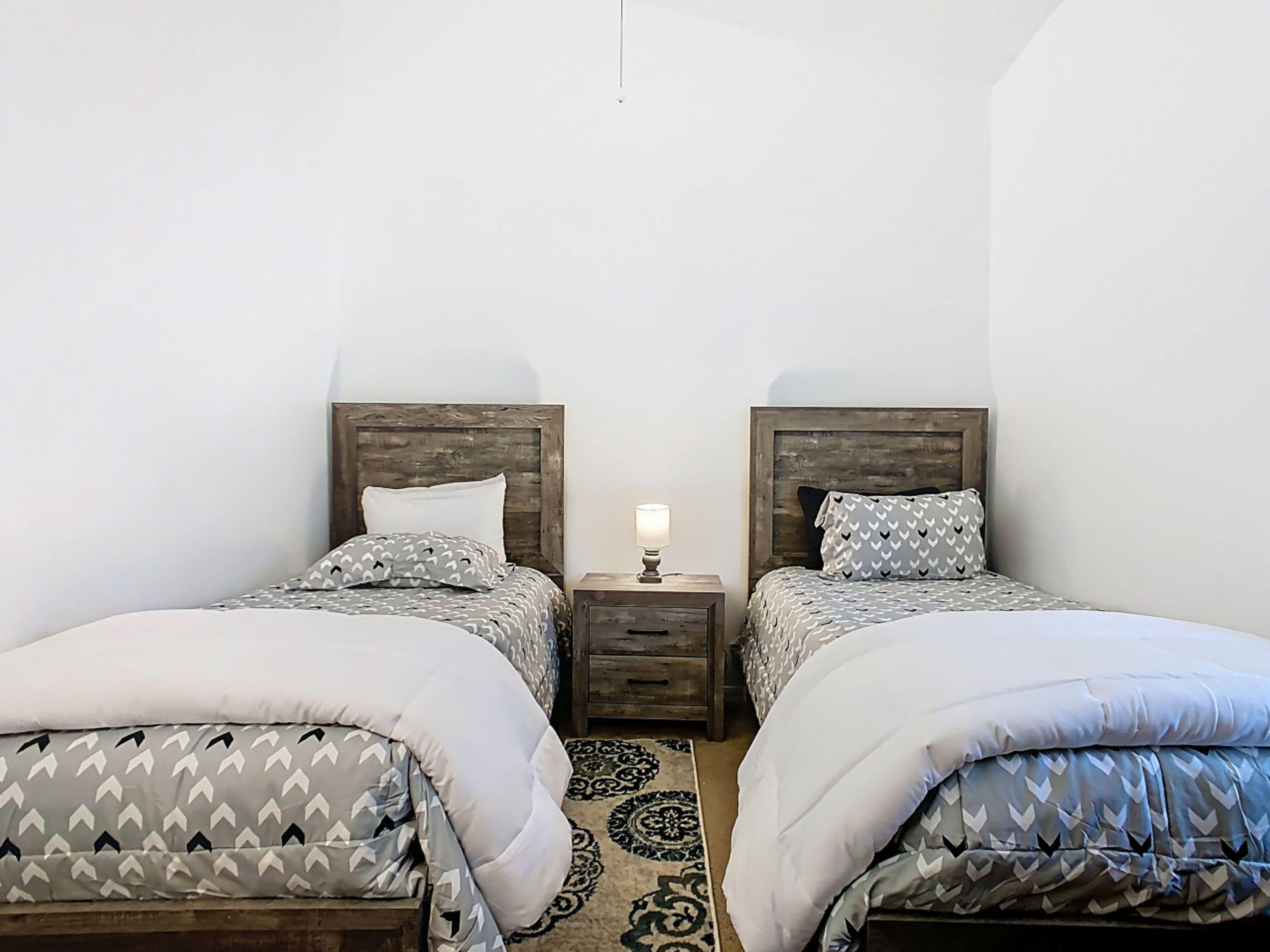 Una habitación con dos camas, cada una con cabecera de madera, una mesita de noche con lámpara y una alfombra estampada en el suelo.