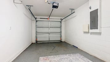 Un garaje con una puerta metálica cerrada, piso de concreto y pared blanca con una ventana.