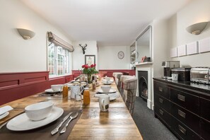 A dining room with a long wooden table set for breakfast, a fireplace, and a kitchen area with a coffee machine. - The Fox at Hurley - Stunning Boutique B B (Maidenhead)