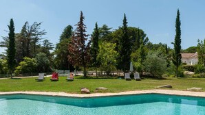 Una piscina con agua cristalina, rodeada de césped, árboles y un edificio blanco al fondo.