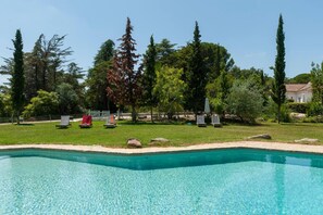 Piscina com água cristalina, rodeada por área gramada com árvores e um edifício branco ao fundo.