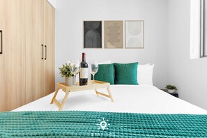 Ein modernes Schlafzimmer mit einem Bett, einem Nachttisch mit Flasche und Gläsern, einer Ablage und einer grünen Decke. An der Wand hängen drei gerahmte Kunstwerke.