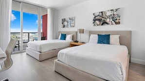 Ein Hotelzimmer mit zwei Betten, einem Schreibtisch und einem Stuhl. An den Wänden hängen zwei Gemälde und das Fenster bietet Blick auf die Stadt.