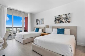 Quarto de hotel com duas camas, uma escrivaninha e uma cadeira. Há duas pinturas na parede e vista para a cidade pela janela.