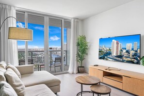 A modern living room with a sofa, a coffee table, a TV, and a view of a cityscape through large windows. - Beach Access Modern Vibe Apt With 1BR in Miami (Hallandale Beach)