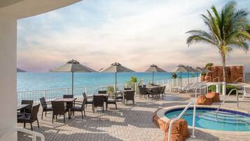 A poolside area with tables and chairs, a pool, and a view of the ocean.