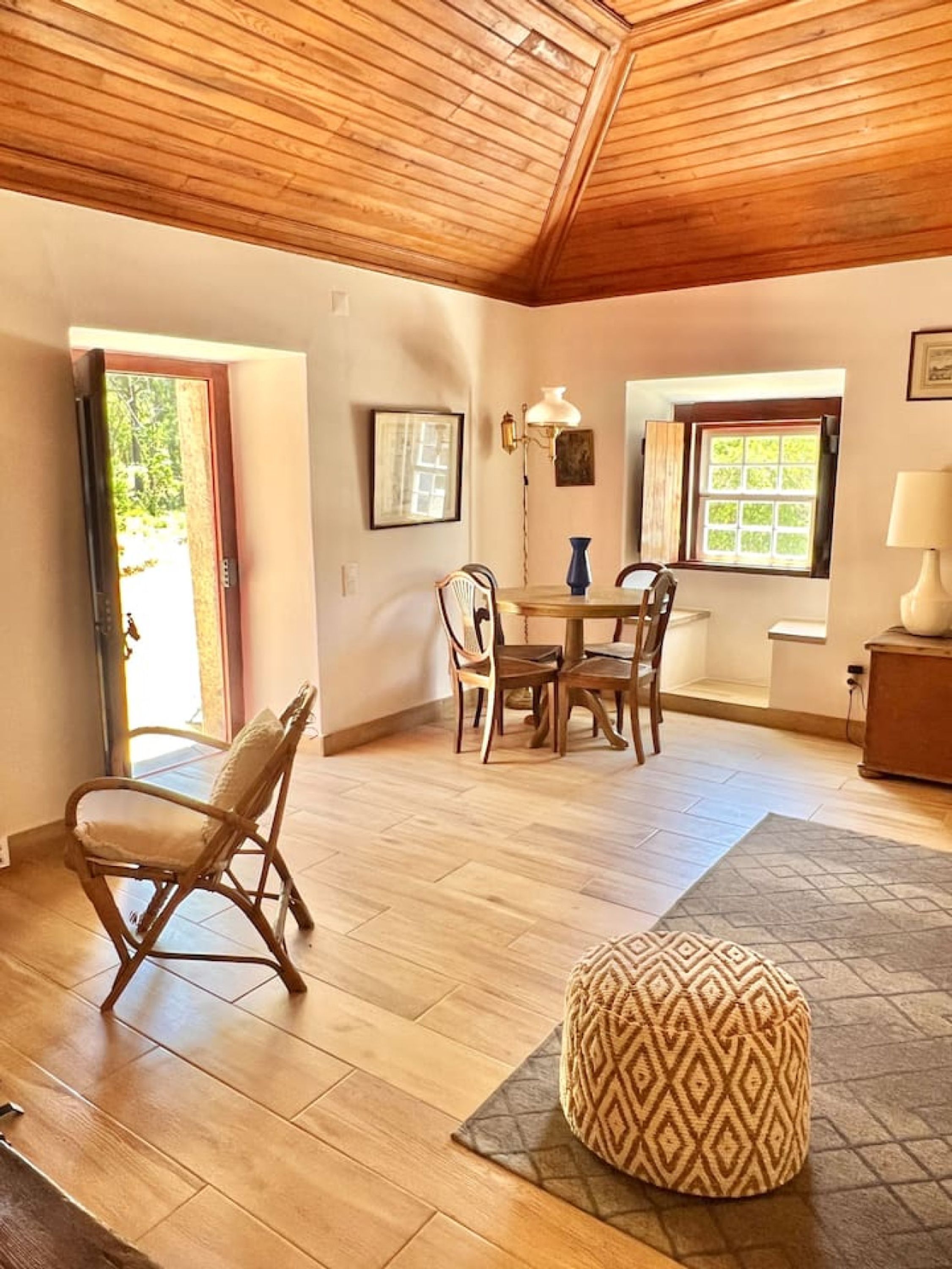 Una habitación con techo de madera, una mesa de comedor con sillas, un otoman decorado y un ventanal con vistas.