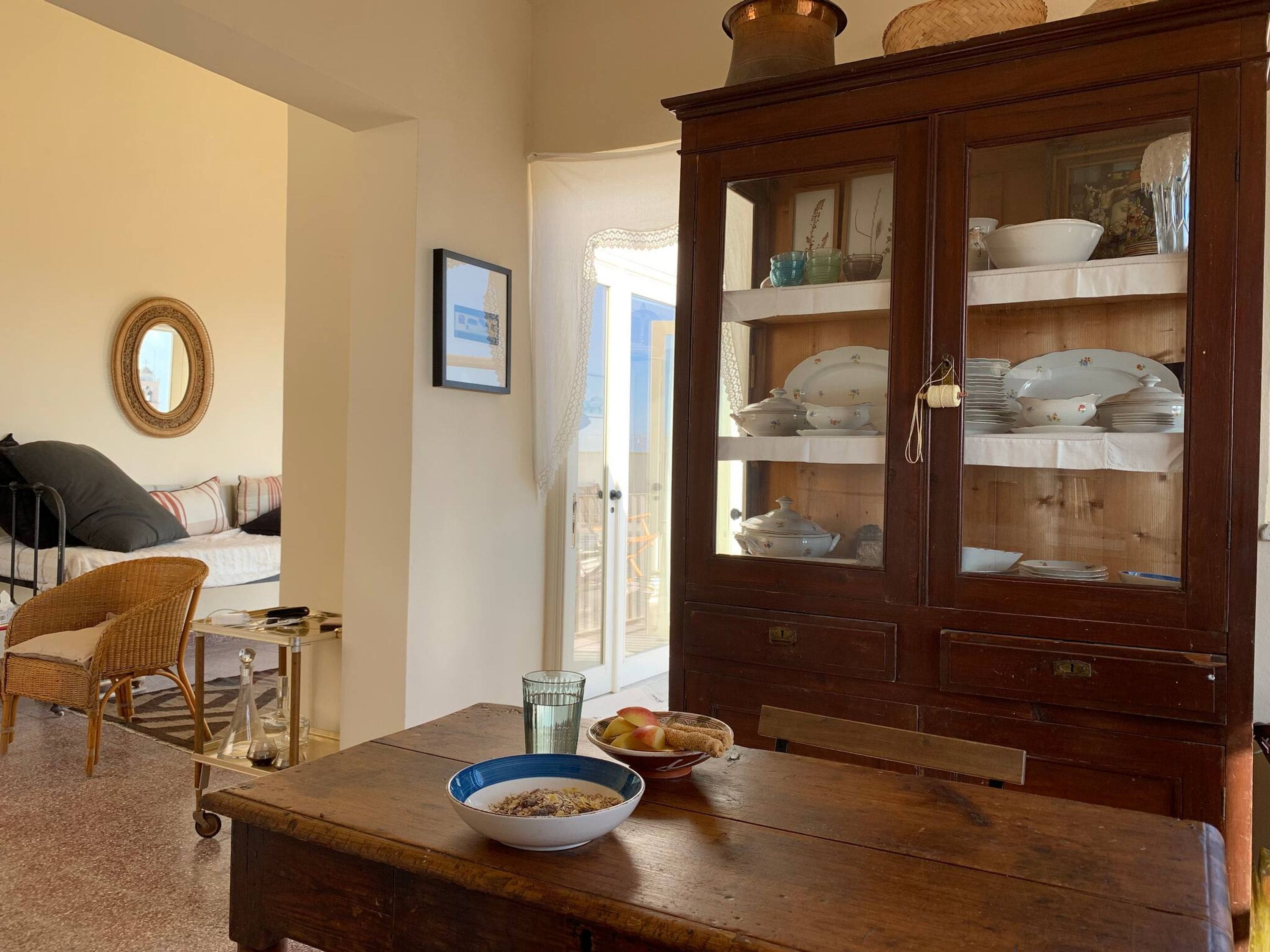 Un comedor con una mesa de madera, sillas de mimbre, un mueble con puertas de vidrio y una cama con almohadas.