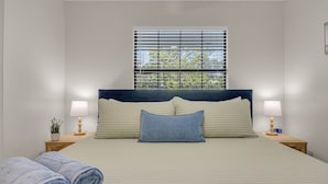 A neatly made bed with a headboard, two bedside lamps, a window with blinds, and a potted plant on the nightstand. - Endless Summer- Beach Pool Firepit 3 King Beds (Ponte Vedra Beach)