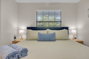 A neatly made bed with a headboard, two bedside lamps, a window with blinds, and a potted plant on the nightstand. - Endless Summer- Beach Pool Firepit 3 King Beds (Ponte Vedra Beach)