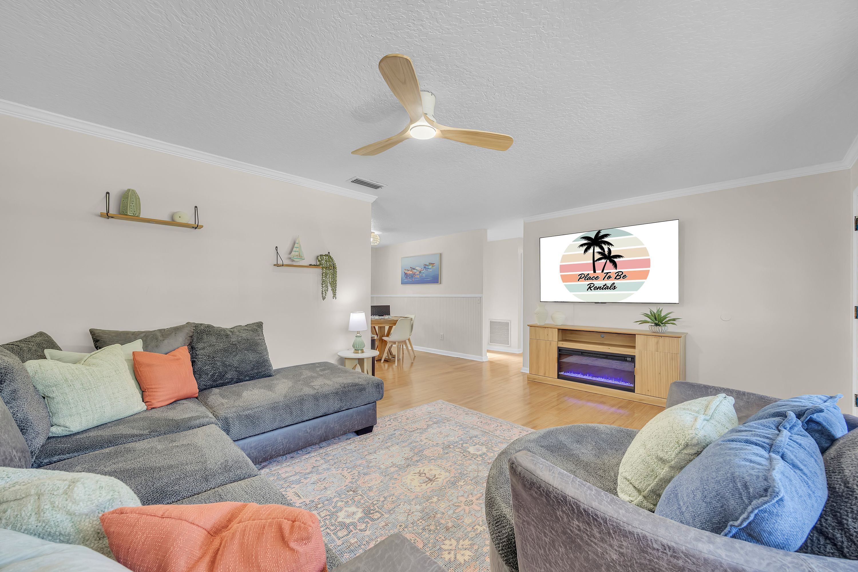 A living room with a sofa, armchairs, a fireplace, a ceiling fan, and a wall-mounted TV displaying 'Place To Be Renios'.