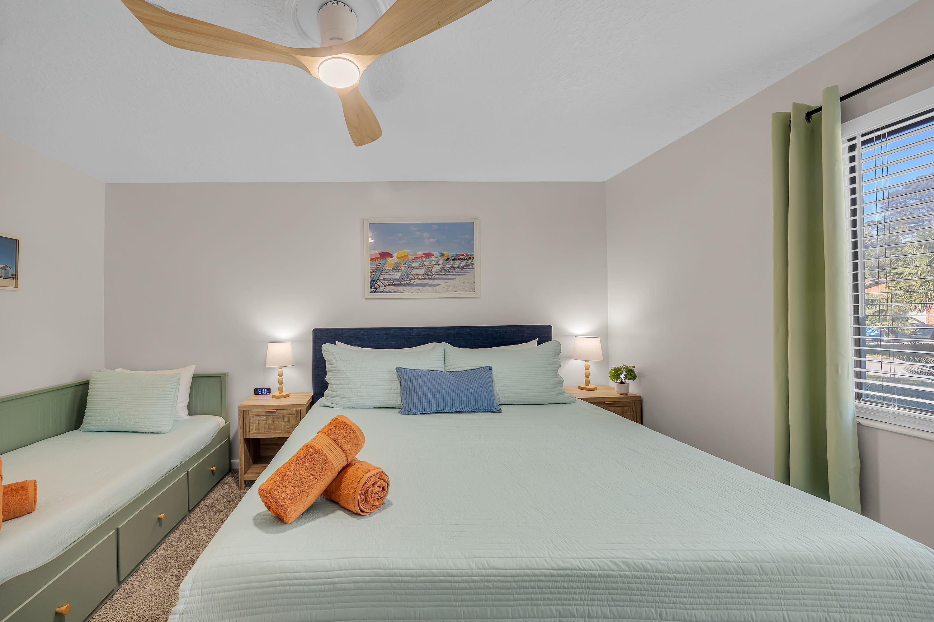 A bedroom with a bed, two bedside tables, a nightstand, a ceiling fan, a window with curtains, and a painting on the wall.