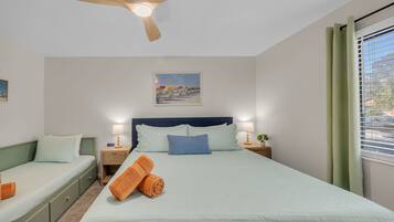 A bedroom with a bed, two bedside tables, a nightstand, a ceiling fan, a window with curtains, and a painting on the wall.