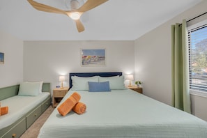 A bedroom with a bed, two bedside tables, a nightstand, a ceiling fan, a window with curtains, and a painting on the wall.
