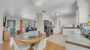 A modern kitchen with a built-in oven, a coffee machine, and a dining area with a round wooden table and white chairs. - Endless Summer- Beach Pool Firepit 3 King Beds (Ponte Vedra Beach)