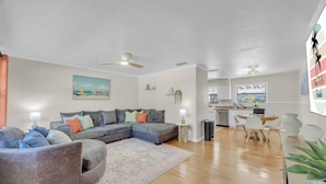 A modern living room with a sectional sofa, a dining area with a table and chairs, a kitchen with white cabinets, and a ceiling fan. - Endless Summer- Beach Pool Firepit 3 King Beds (Ponte Vedra Beach)