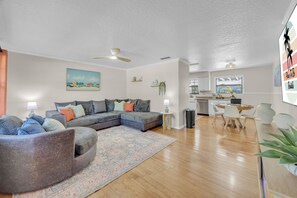 A modern living room with a sectional sofa, a dining area with a table and chairs, a kitchen with white cabinets, and a ceiling fan. - Endless Summer- Beach Pool Firepit 3 King Beds (Ponte Vedra Beach)