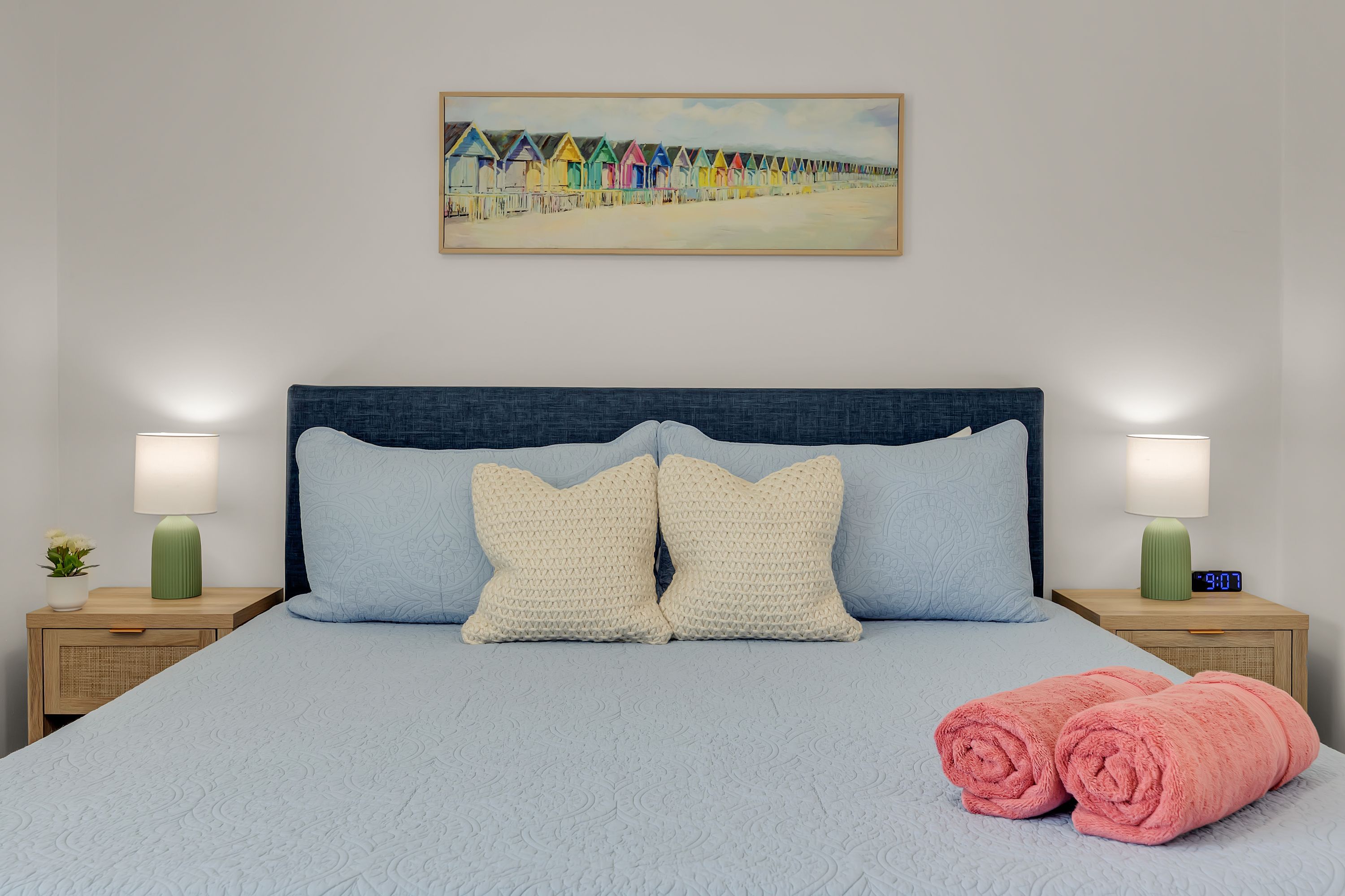 A neatly made bed with a blue headboard, two pillows, a pair of rolled towels, and a painting of colorful beach huts on the wall.