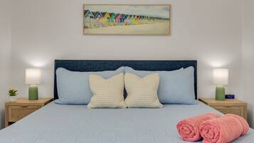 A neatly made bed with a blue headboard, two pillows, a pair of rolled towels, and a painting of colorful beach huts on the wall.
