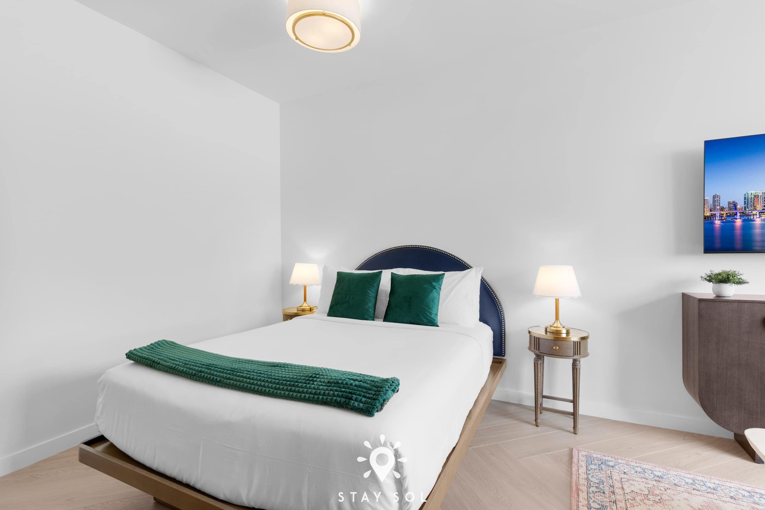 Ein Hotelzimmer mit einem Bett, Nachttischlampen, einem Nachttisch, einer kleinen Kommode und einem Bild an der Wand.