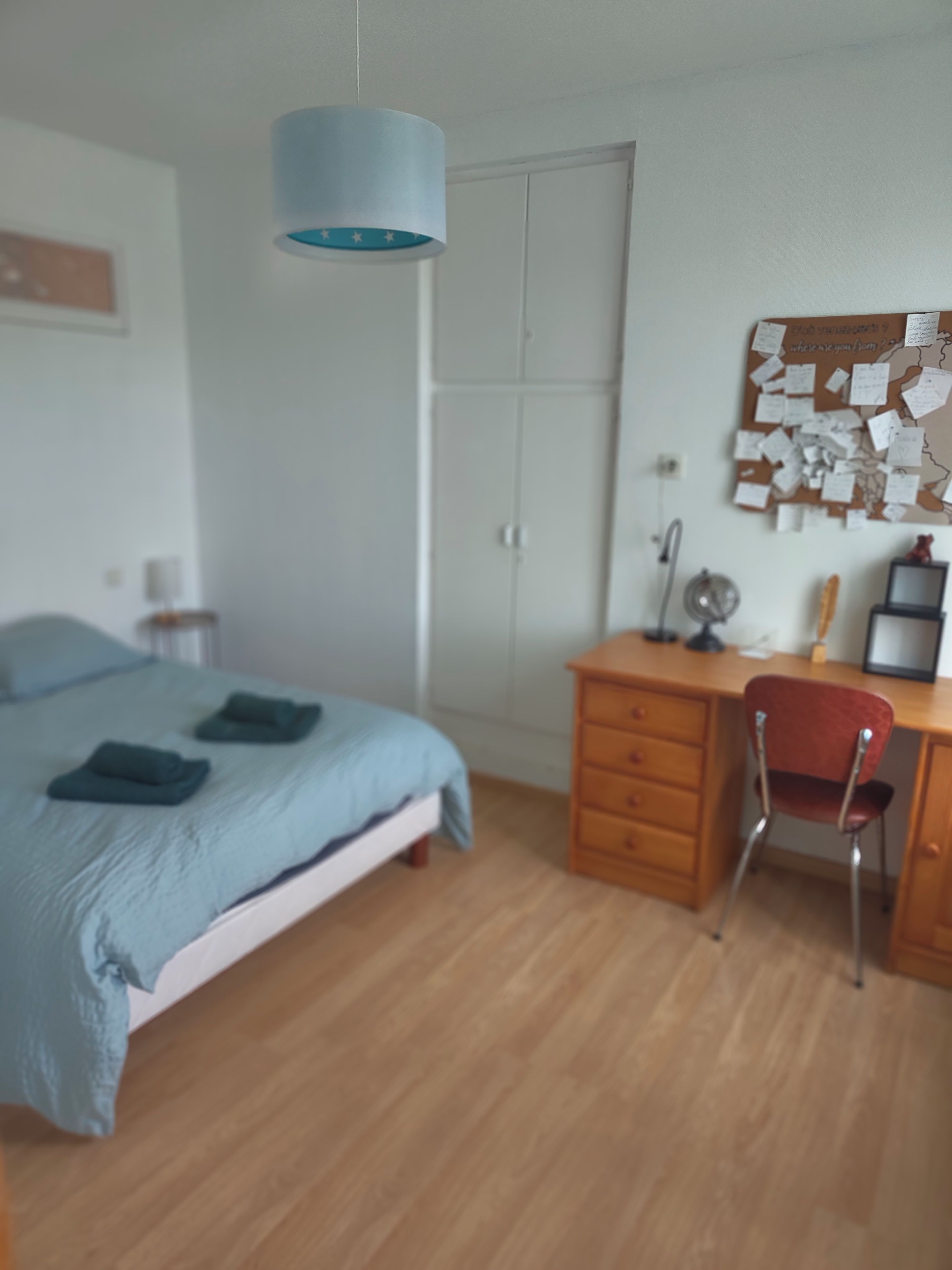 Een slaapkamer met een bed, een bureau, een stoel, een kledingkast en een prikbord met notities.
