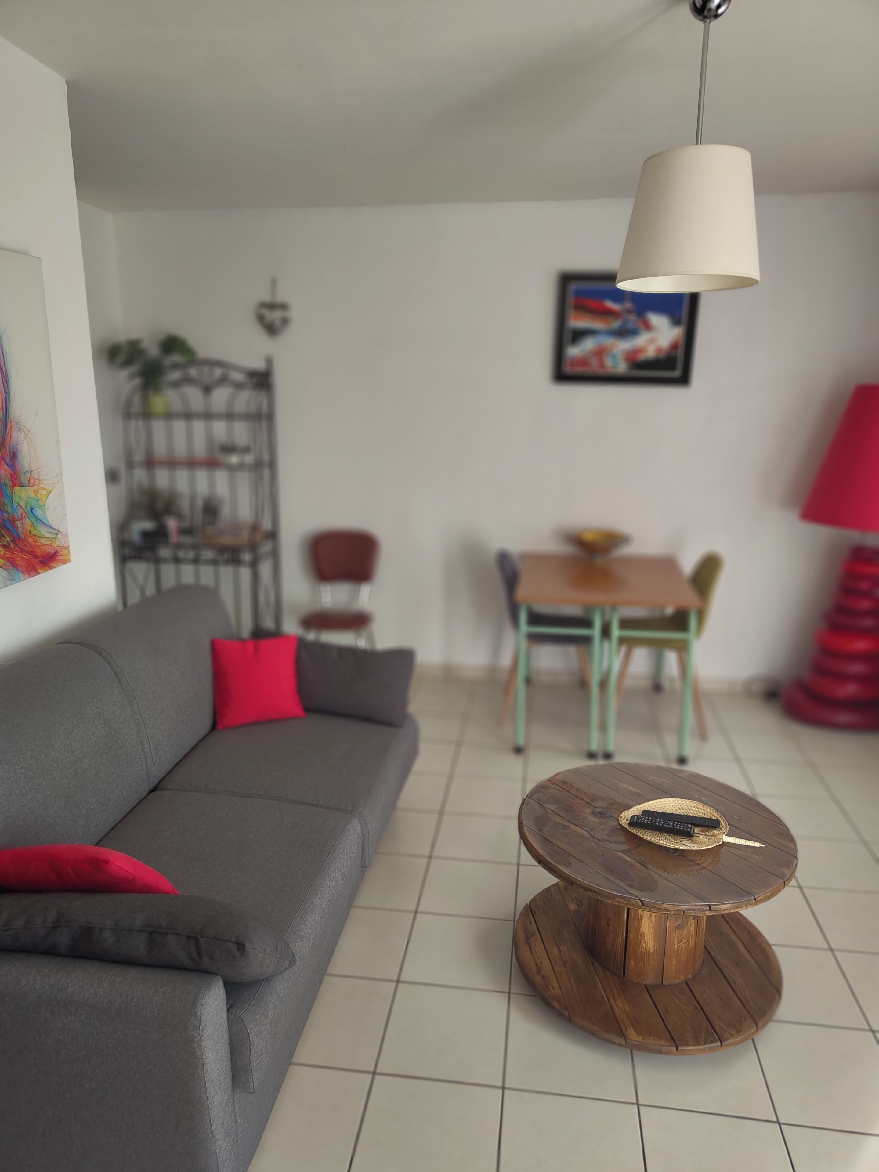 A living room with a grey sofa, a wooden coffee table, a dining table with chairs, a hanging lamp, and a painting on the wall.