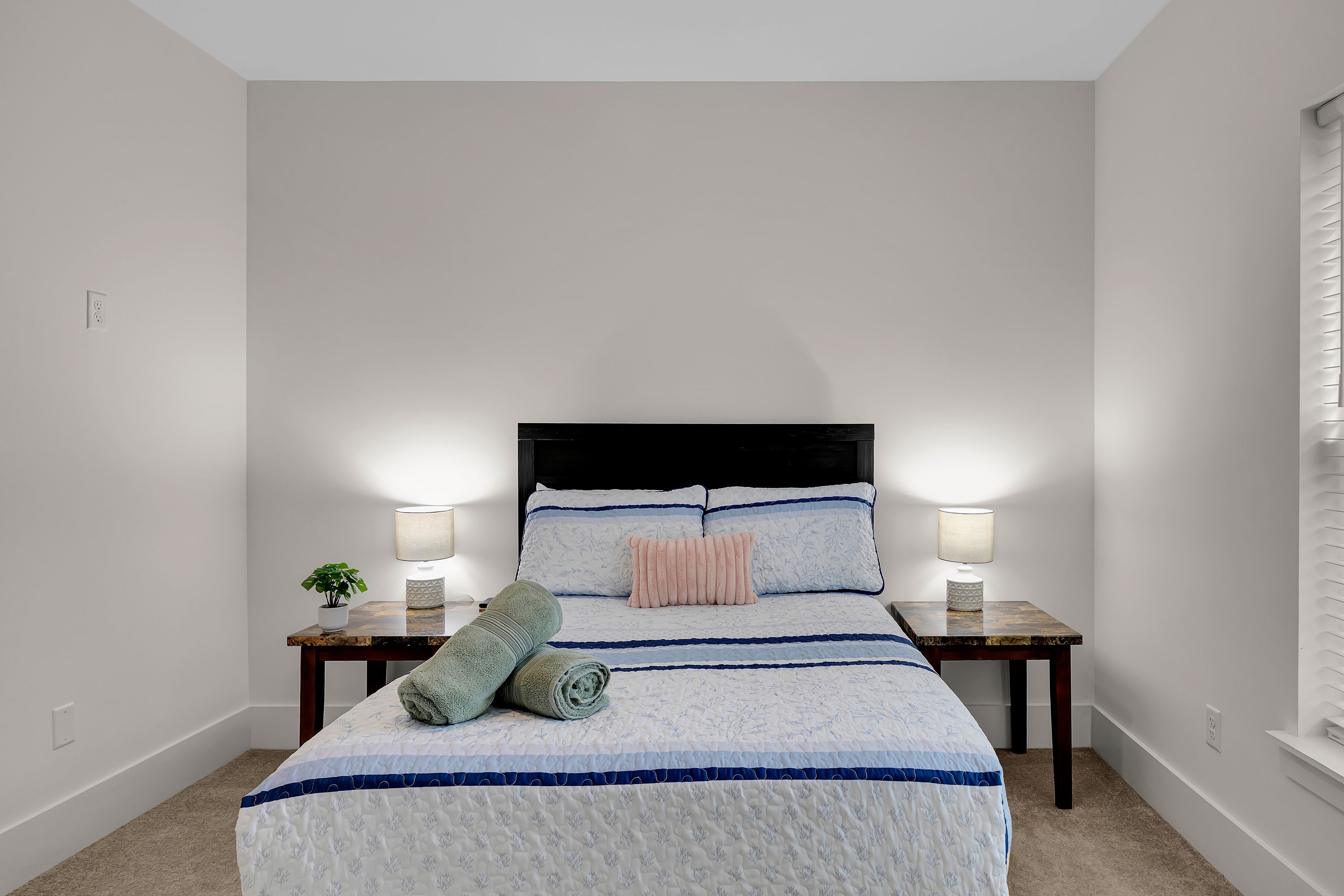Un dormitorio con una cama, dos mesitas de noche, una lámpara en cada mesita y una planta en maceta, además de una ventana con persianas.