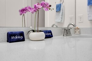 Un baño con un lavamanos blanco, un espejo y un florero con flores moradas. Hay dos toallas con la palabra 'Makeup'.