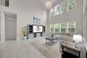 A spacious living room with a large TV, a sofa, a coffee table, and a chandelier. - Salt Air 6BR Game Room Walk to Beach Luxe (Ponte Vedra Beach)