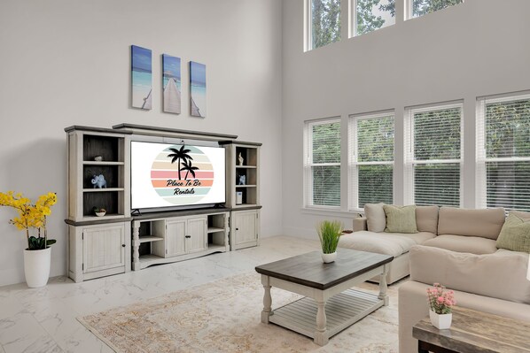 A spacious living room with a large flat-screen TV, a beige sofa, a coffee table, and a decorative rug.