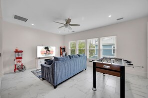 A spacious living room with a sofa, a foosball table, a ceiling fan, and a large window with blinds.